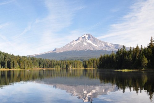 A serene lake reflects a snow-capped mountain surrounded by lush green forests under a clear blue sky.