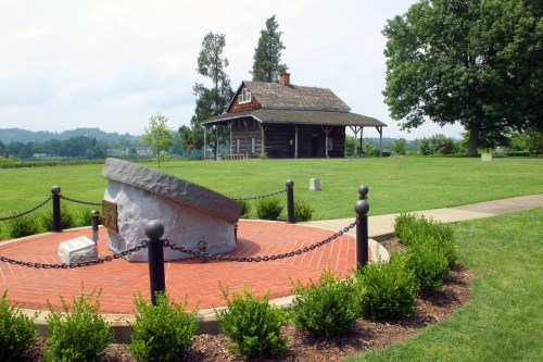A historic log cabin surrounded by greenery, with a stone monument in the foreground and hills in the background.