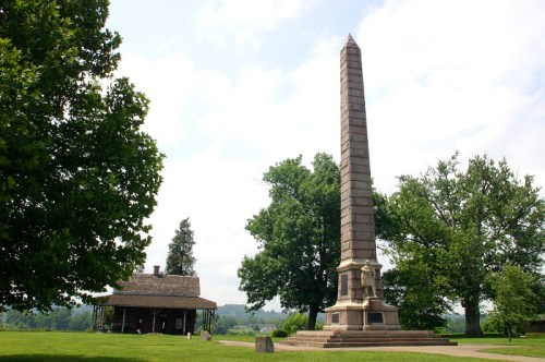 A tall stone obelisk stands beside a small historic building, surrounded by lush green trees and a cloudy sky.