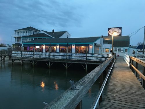 A waterfront restaurant at dusk, with a wooden pier leading to it and a sign illuminated above.