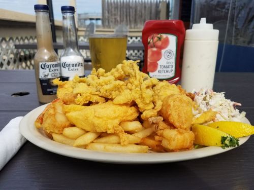 A plate of fried seafood and fries, accompanied by coleslaw, ketchup, and two bottles of hot sauce, with a drink in the background.