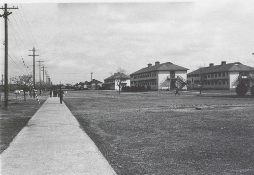 Black and white photo of a wide street with sidewalks, buildings in the background, and people walking.