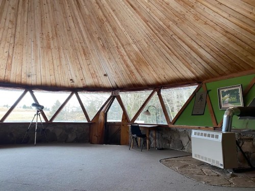 Interior of a circular room with a wooden ceiling, stone walls, a telescope, and a table by large windows.