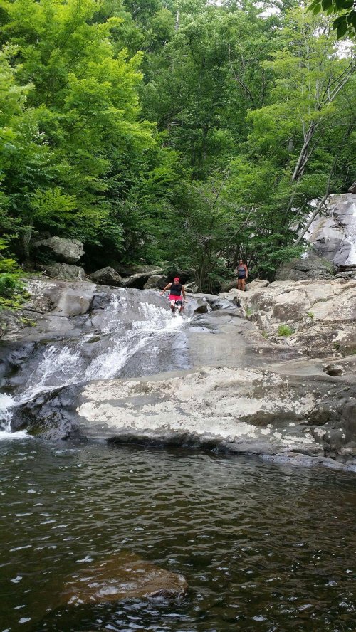 Two people explore a rocky waterfall surrounded by lush green trees and a calm pool of water below.
