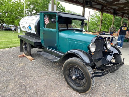A vintage teal truck with a white tank in the back, parked under a shelter at an outdoor event.