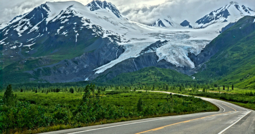 Winding road through lush green landscape with snow-capped mountains and a glacier in the background.