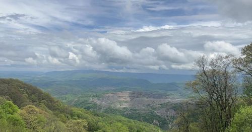A panoramic view of lush green mountains under a cloudy sky, with a valley and a mining site visible in the distance.