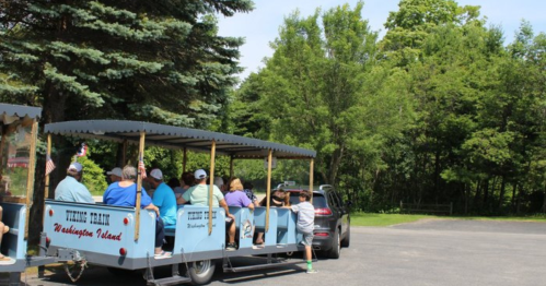A blue train ride with passengers is parked beside a car in a green, wooded area on a sunny day.