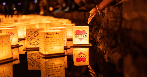 Hands reaching towards illuminated lanterns floating on water, each adorned with colorful drawings and messages.