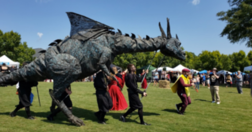 A large dragon puppet is paraded by performers in costumes at an outdoor festival, with tents in the background.