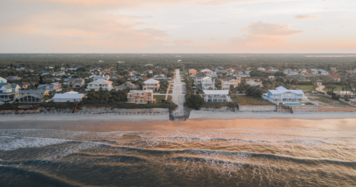 Aerial view of a coastal neighborhood at sunset, with houses lining the beach and gentle waves lapping at the shore.