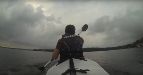 A person kayaking on a calm lake under a cloudy sky, paddling away from the camera.