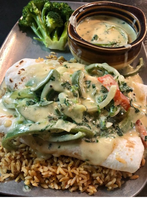 A plate with a burrito topped with green peppers and creamy sauce, served with rice and steamed broccoli on the side.