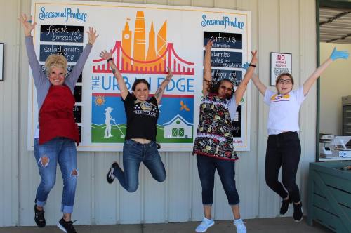 Four women jump joyfully in front of a colorful market sign, showcasing fresh produce and seasonal offerings.