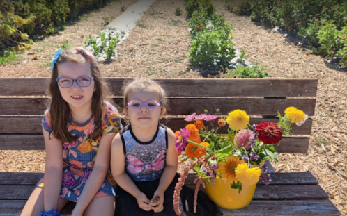 Two girls sit on a bench in a garden, surrounded by colorful flowers in a yellow bucket.