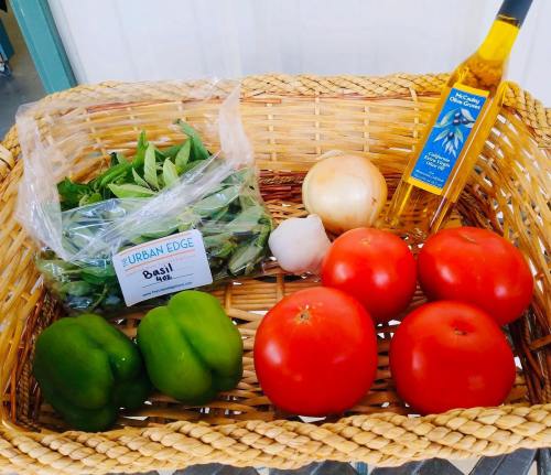 A woven basket filled with fresh basil, green peppers, tomatoes, an onion, garlic, and a bottle of olive oil.