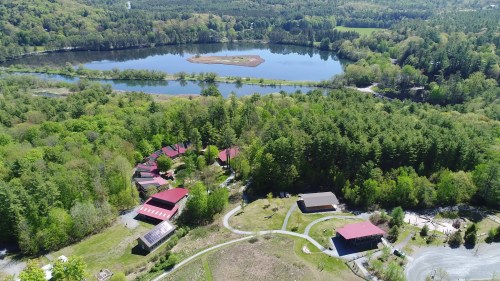 Aerial view of a lush green landscape with a lake, surrounded by trees and several buildings with red roofs.