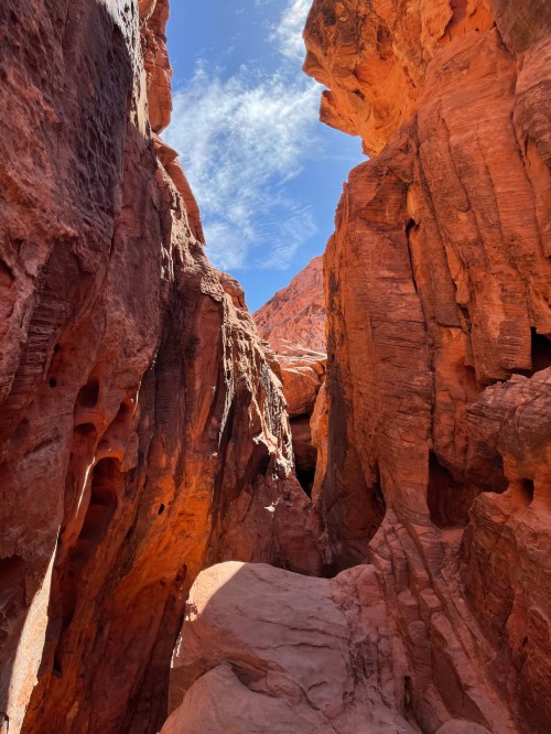 Narrow canyon walls of red rock under a blue sky with scattered clouds. Sunlight illuminates the rugged terrain.