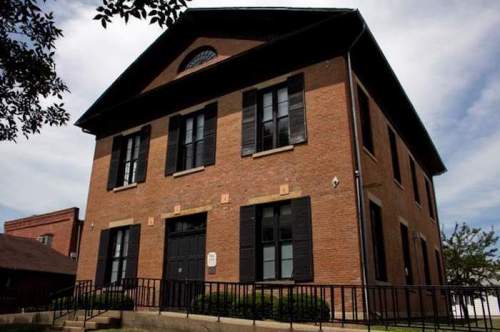 Historic brick building with large windows and a sloped roof, surrounded by greenery under a cloudy sky.