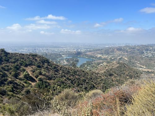 A panoramic view of a cityscape with hills and a lake under a partly cloudy sky.