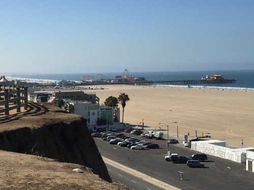 A view of a sandy beach with a pier and amusement park in the distance, framed by palm trees and parked cars.