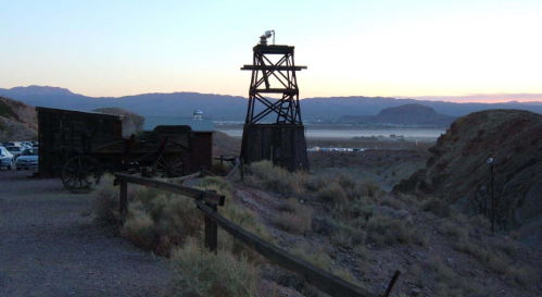 A wooden mining tower stands on a hillside at sunset, overlooking a valley with distant mountains and parked cars.