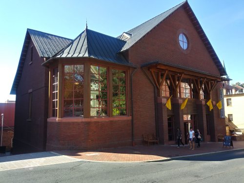 A brick building with large windows and a peaked roof, featuring a welcoming entrance and people walking by.