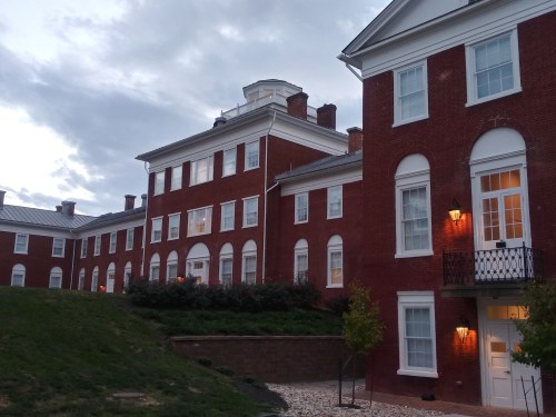 A red brick building with white trim, featuring multiple windows and a grassy area in front, under a cloudy sky.