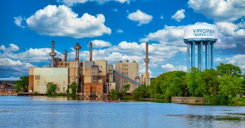 Industrial facility by a lake with a water tower labeled "Virginia Queen City" under a blue sky with clouds.