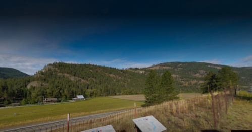 A scenic view of rolling hills and fields under a blue sky, with trees and a fence in the foreground.