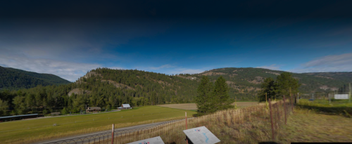 A scenic view of rolling hills and fields under a blue sky, with trees and a fence in the foreground.