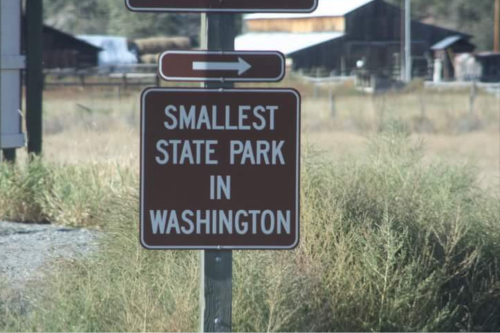 Sign indicating "Smallest State Park in Washington" with an arrow pointing left, surrounded by grass and buildings in the background.