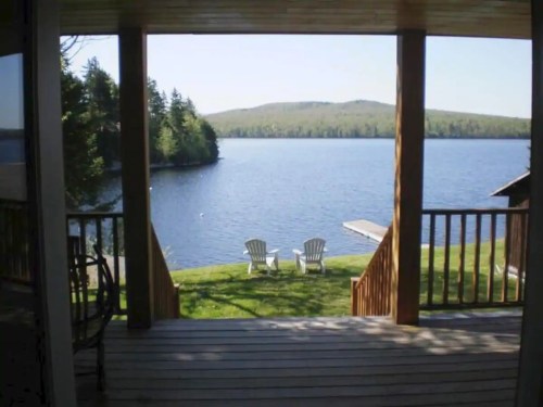 View from a porch overlooking a serene lake, with two chairs facing the water and lush greenery in the background.