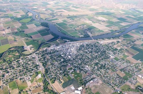 Aerial view of a small town surrounded by farmland and a winding river, with green fields and mountains in the background.