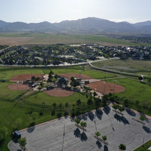 Aerial view of a baseball field surrounded by green grass, mountains, and a residential area in the background.