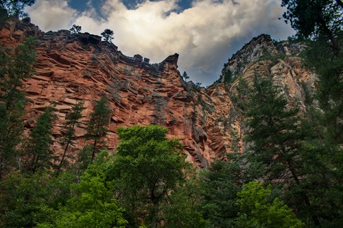 A towering red rock cliff surrounded by lush green trees under a cloudy sky.
