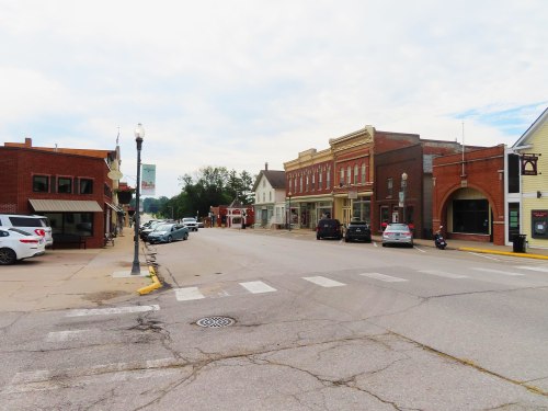 A quiet street in a small town featuring brick buildings, parked cars, and a cloudy sky.