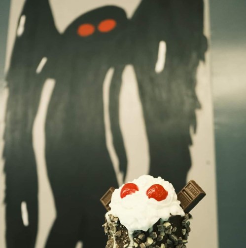 A dessert topped with whipped cream and cherries in front of a dark, abstract wall painting with red eyes.