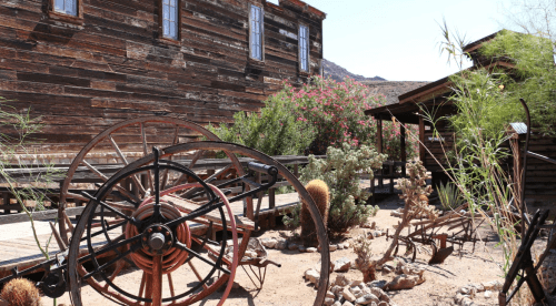 A rustic scene featuring a large wagon wheel, desert plants, and weathered wooden buildings under a clear blue sky.