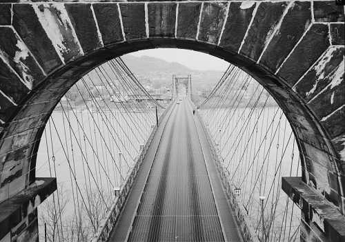 Black and white image of a suspension bridge viewed through a stone arch, with a river and distant hills in the background.