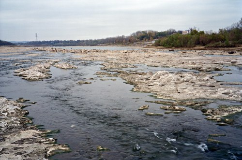 A calm river flows over rocky terrain, surrounded by trees and a cloudy sky in the background.