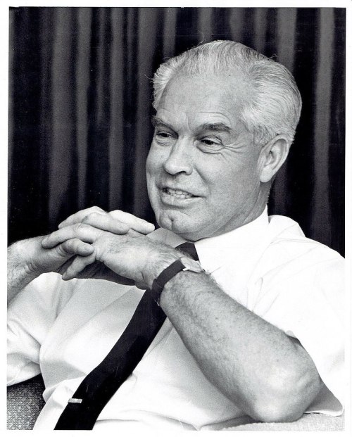 A black and white portrait of a smiling man in a white shirt and tie, sitting with hands clasped.