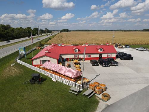 Aerial view of a red-roofed market with pumpkins outside, near a highway and cornfields under a blue sky.