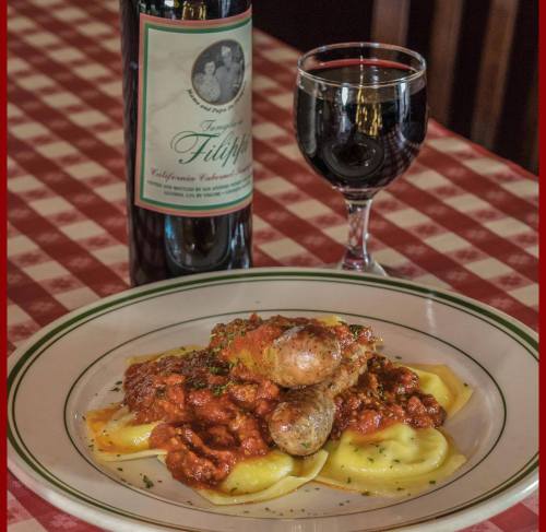 A plate of ravioli topped with meat sauce and sausages, accompanied by a glass of red wine and a bottle on a checkered tablecloth.