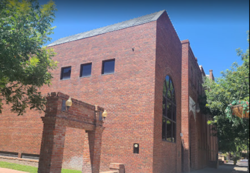 A brick building with large windows and an archway, surrounded by trees under a clear blue sky.