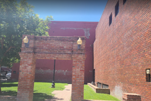 A brick archway leads to a grassy area, with a red brick wall and trees in the background under bright sunlight.