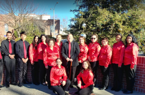 A group of musicians in red shirts and black pants pose together outdoors, smiling in front of a decorated tree.