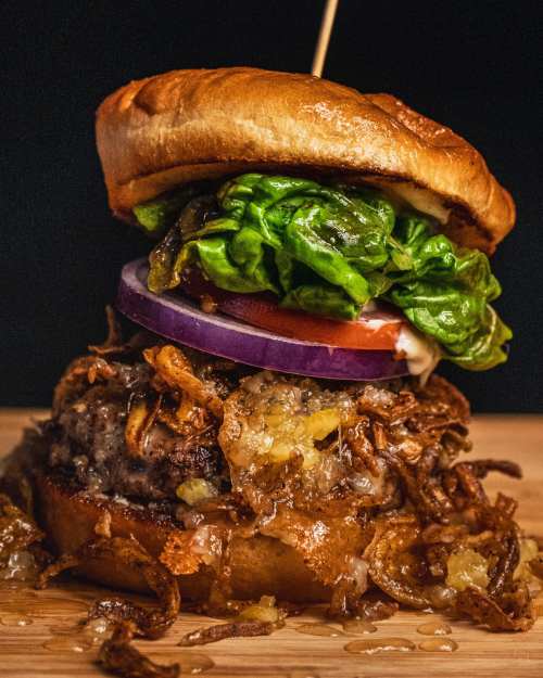 A close-up of a juicy burger stacked with lettuce, tomato, onion, and crispy toppings on a toasted bun.