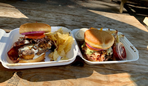 Two burgers on a tray: one with bacon and cheese, the other with toppings, served with chips and dipping sauces.