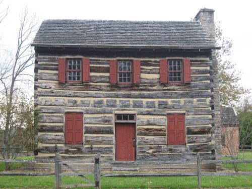 A rustic log cabin with red shutters and a front door, surrounded by greenery and trees.